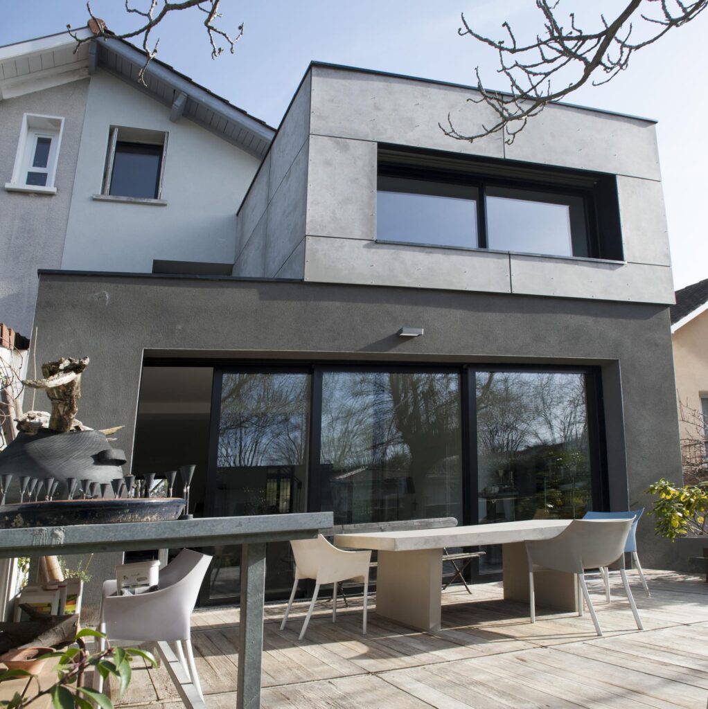 Extension moderne façade bardage gris et terrasse bois. Extension contemporaine à façade grise (ciment et enduit) sur maison ancienne, ouvrant sur une terrasse bois avec table et chaises design.