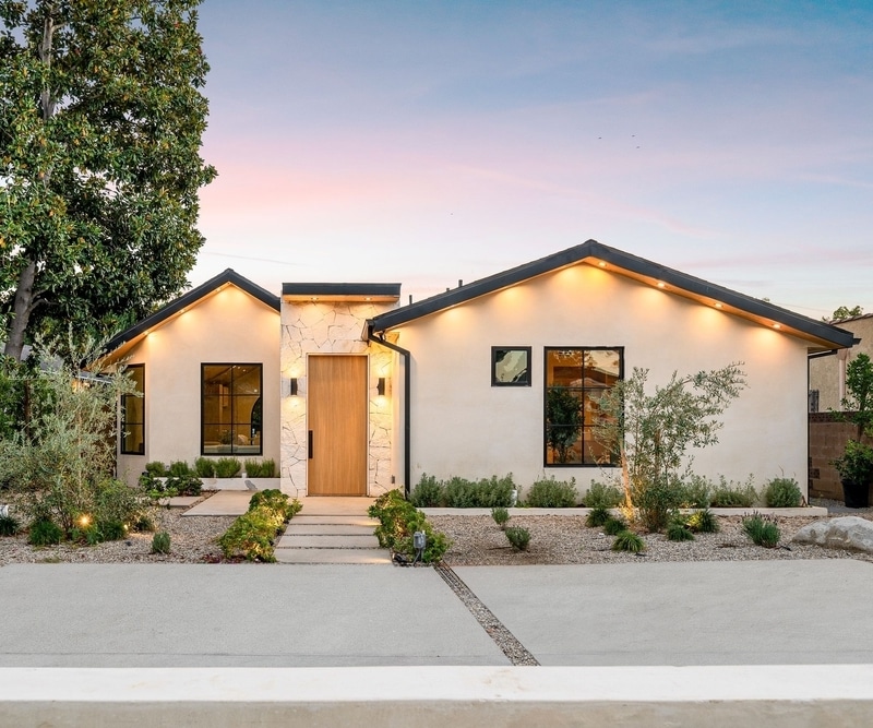Maison moderne : Façade élégante au crépuscule, lumière chaude. Façade de maison moderne au crépuscule. Murs clairs, toit noir, porte en bois et mur d'entrée en pierre naturelle. Éclairage soffite chaleureux.