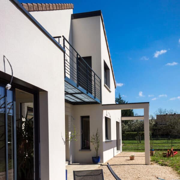 Maison moderne : Façade blanche avec balcon en métal et terrasse jardin. Maison contemporaine blanche, balcon grillagé noir et pergola. Terrasse en gravier menant à un grand jardin sous ciel bleu.