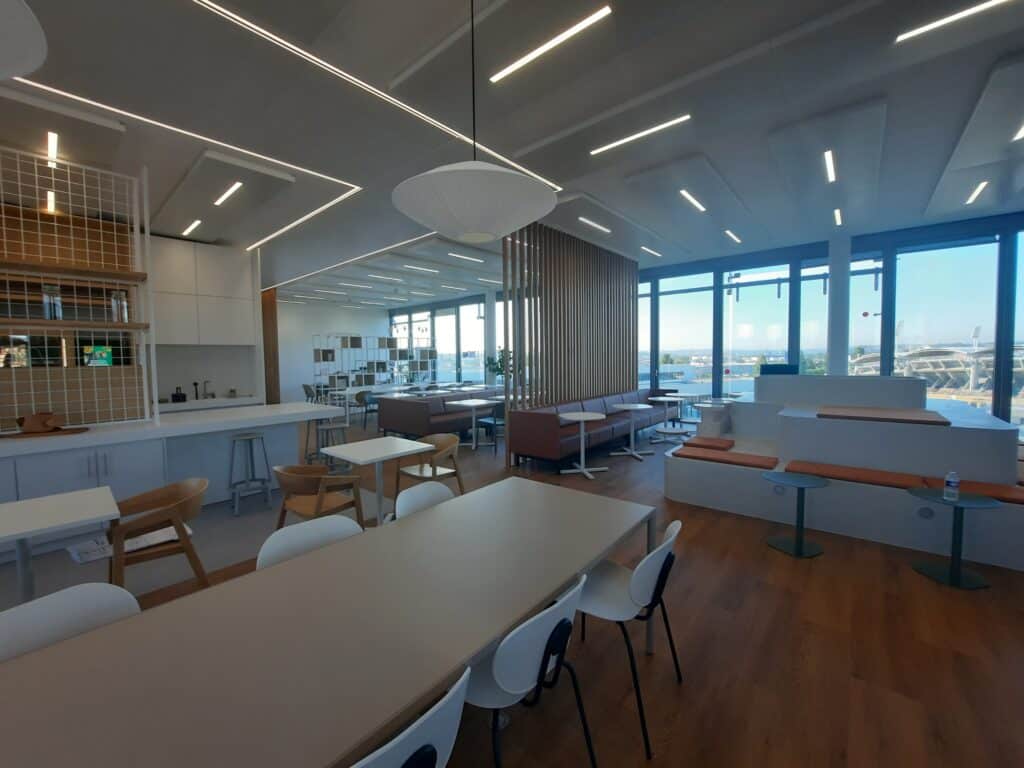 Espace de Coworking Moderne : Aménagement Bureau et Café Espace café d'entreprise moderne avec cuisine, tables design, banquettes et grandes baies vitrées avec vue urbaine.